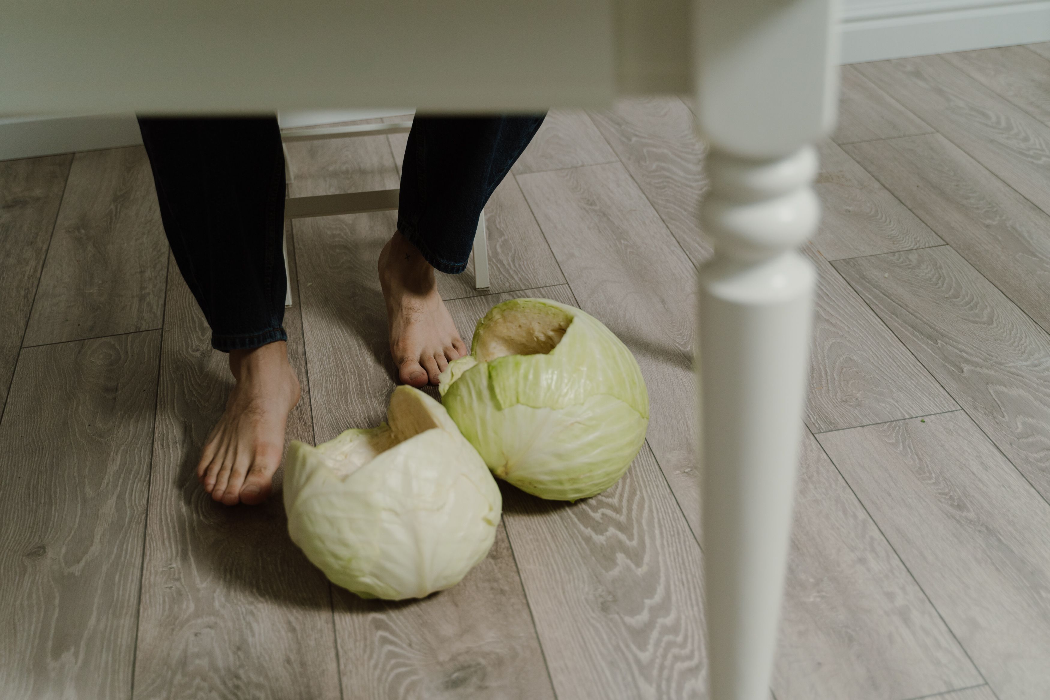 Close-up of bare feet stepping next to cabbages on wooden floor indoors.