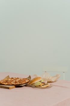 A minimalist still life of a burger and pizza on a pastel table.