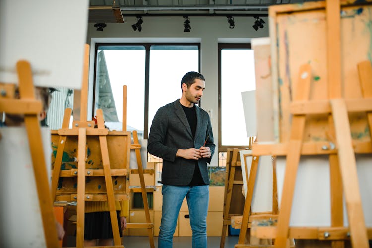 Man In Black Blazer Standing In A Room Of Wooden Easels
