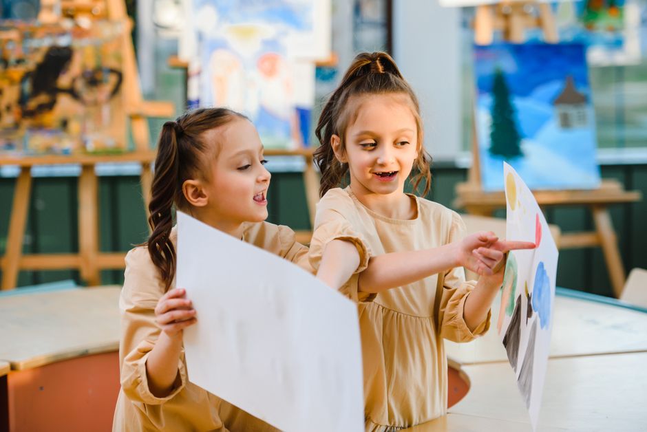 Two young girls happily sharing and discussing their paintings in an art classroom.
