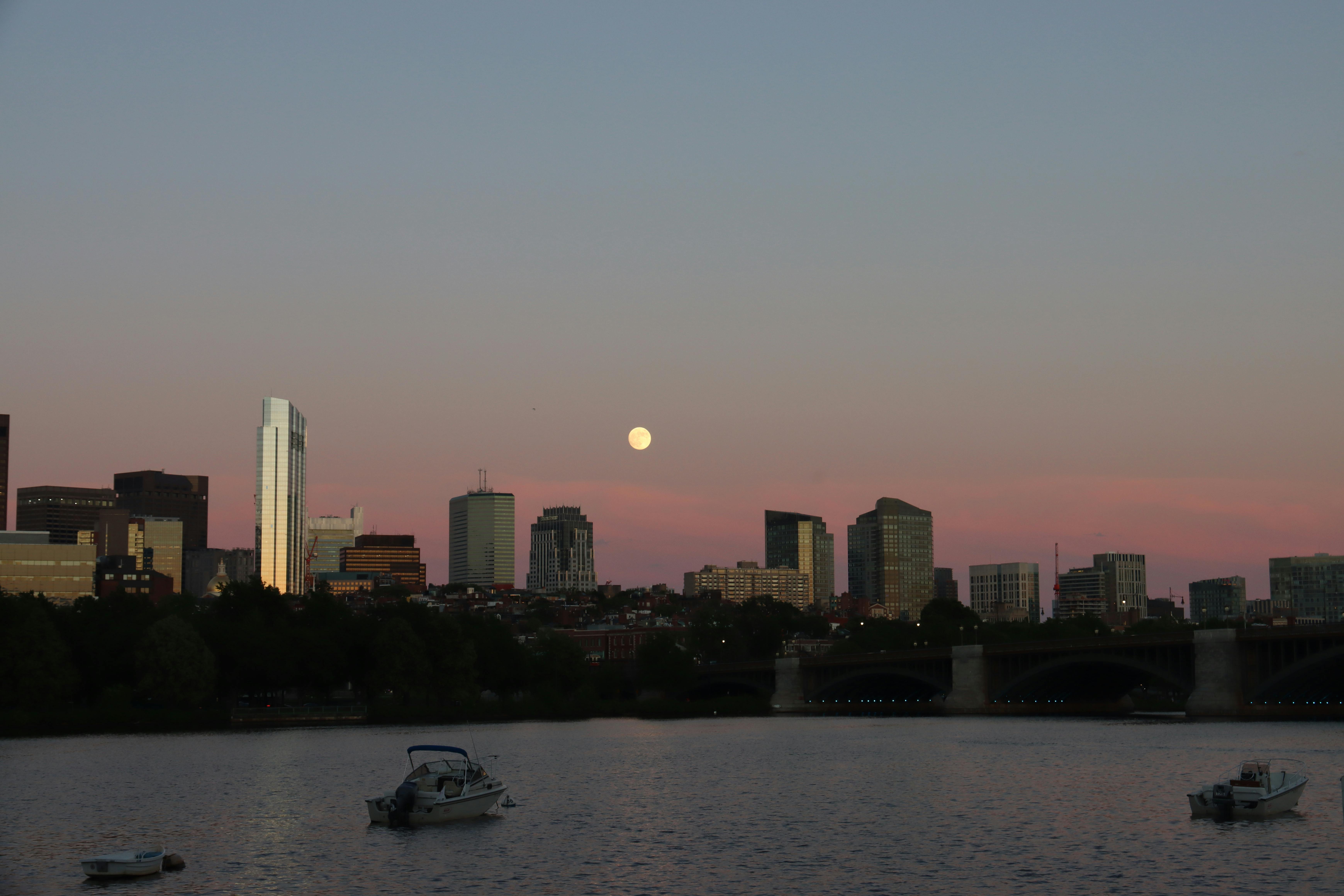Scenic view of Boston skyline and Charles River at dusk with a full moon backdrop.