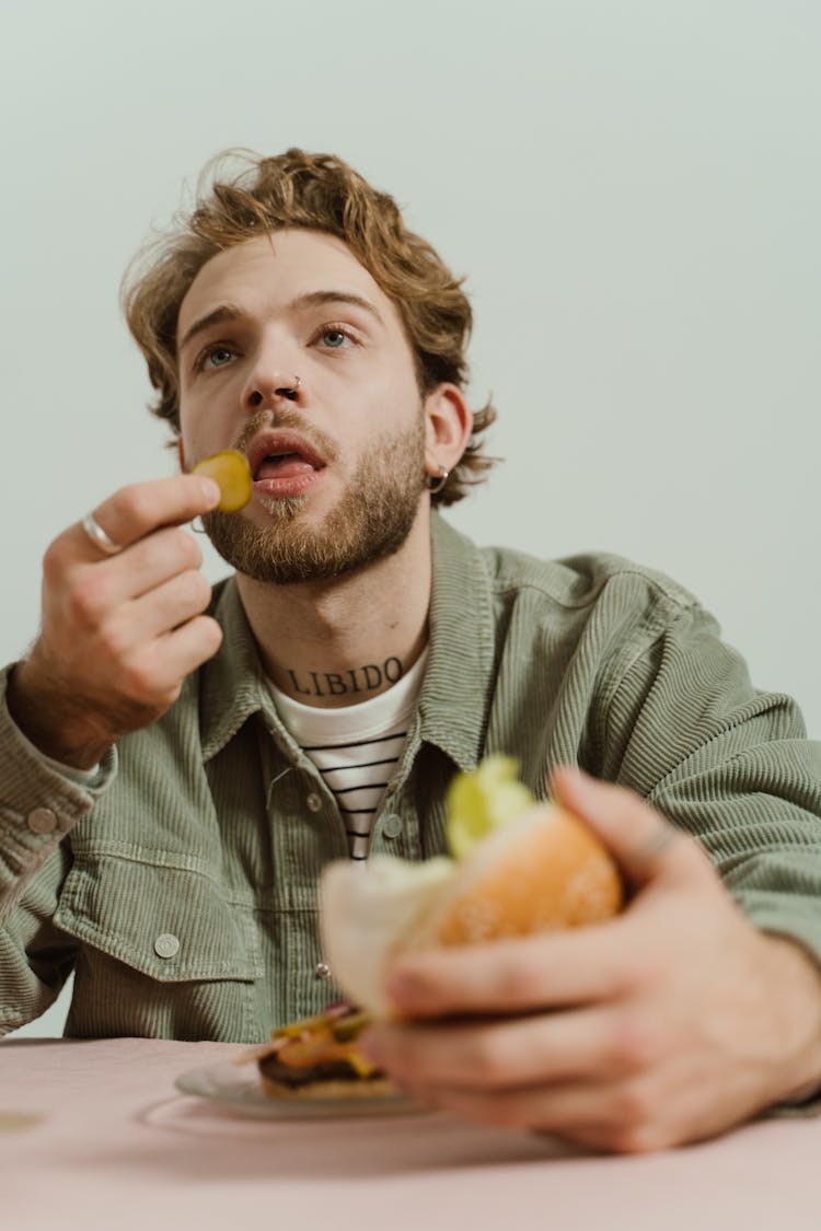 Man In Green Jacket Eating Sliced Pickle While Holding The Bun Of Hamburger