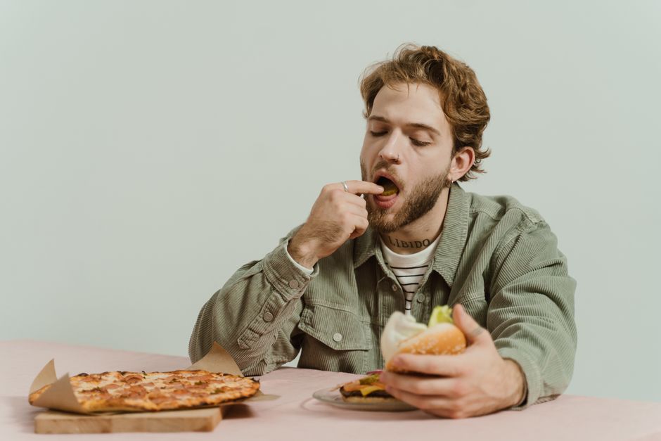 室内に座る大人の男性がリラックスした表情でピザとハンバーガーを食べています。