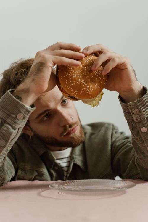 Free A thoughtful man holding a burger inside, sitting at a table with a plate. Stock Photo