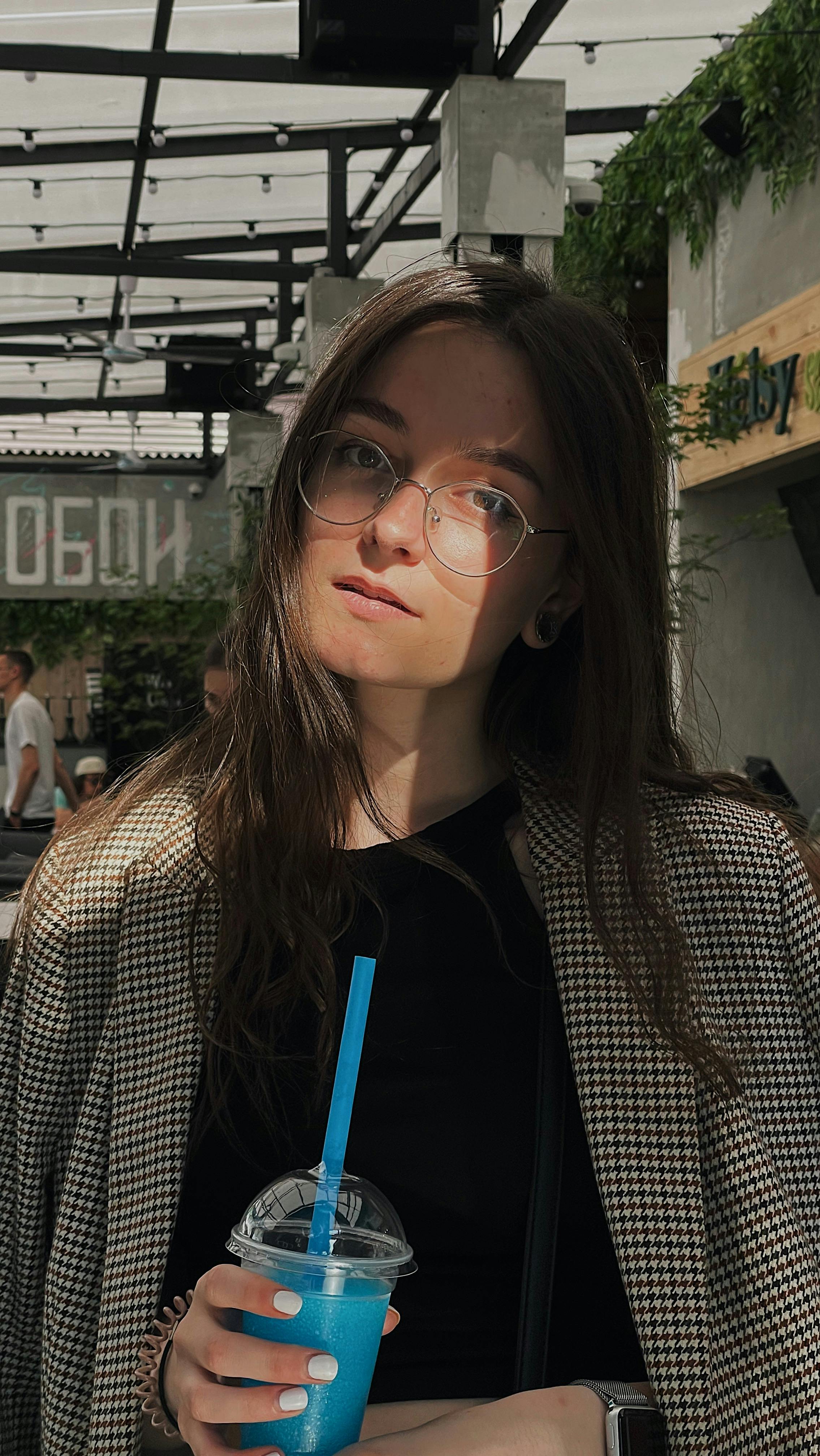 A young woman with eyeglasses enjoys a refreshing blue drink while standing outdoors in a modern setting.