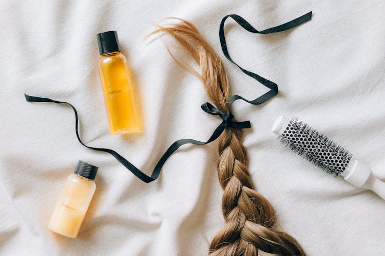 A Braided Hair With Ribbon On White Textile Beside A Brush And Bottles