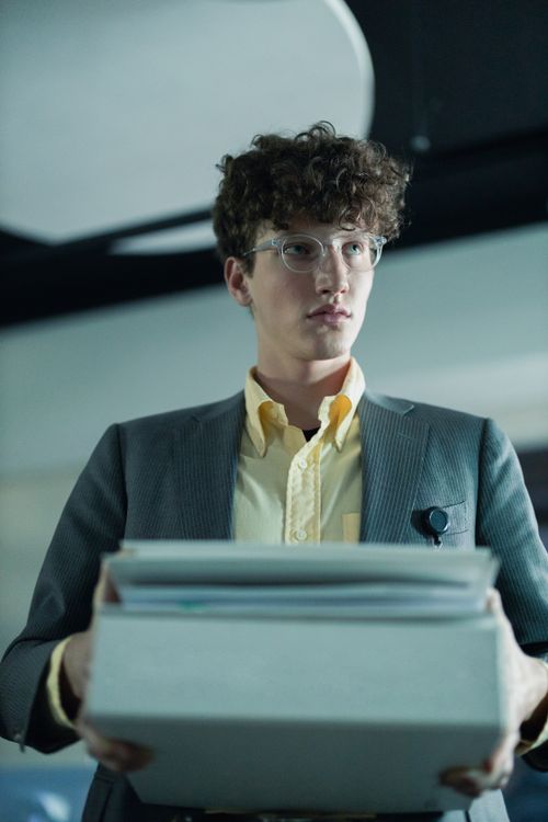 Free Man in glasses, stylish gray blazer, and yellow shirt holding documents in a modern office setting. Stock Photo
