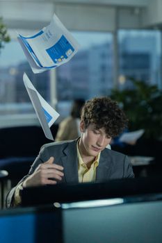 A young man in a gray jacket tosses documents in a modern office setting, creating a dynamic business scene.