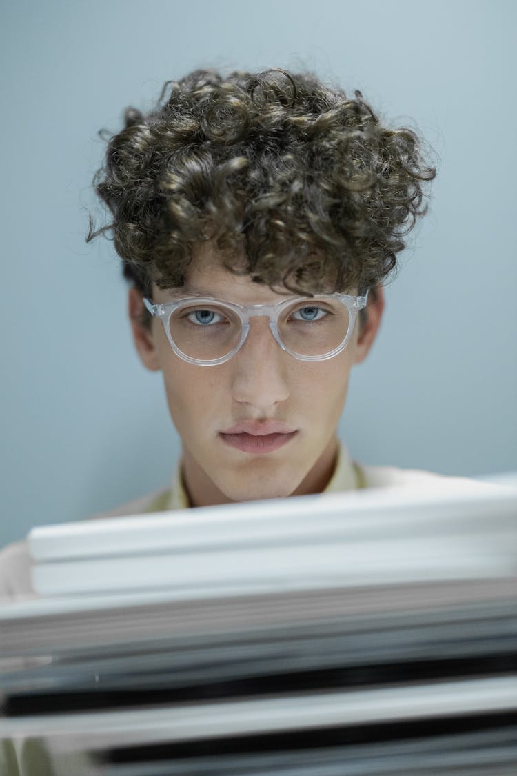 Man In White Framed Eyeglasses With Curly Hair Sitting Behind A Pile Of Books