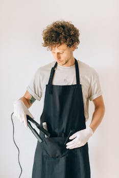 A male hairdresser in a salon holding a hair iron, dressed in a black apron.