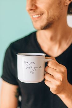 A close-up shot of a smiling man holding a white mug with an encouraging message.