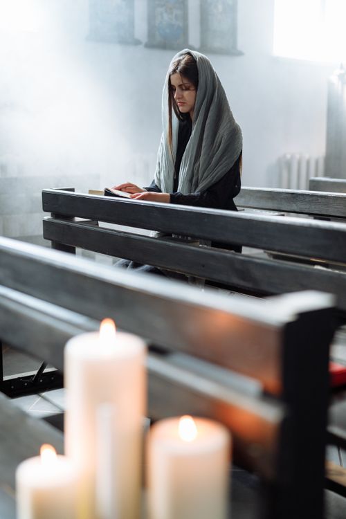 Free A contemplative woman in a headscarf sitting in a church pew with candles in the foreground. Stock Photo