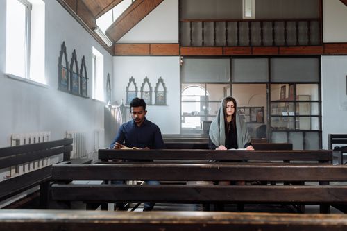 Free Two individuals seated in a serene church setting, absorbed in quiet reflection and reading. Stock Photo