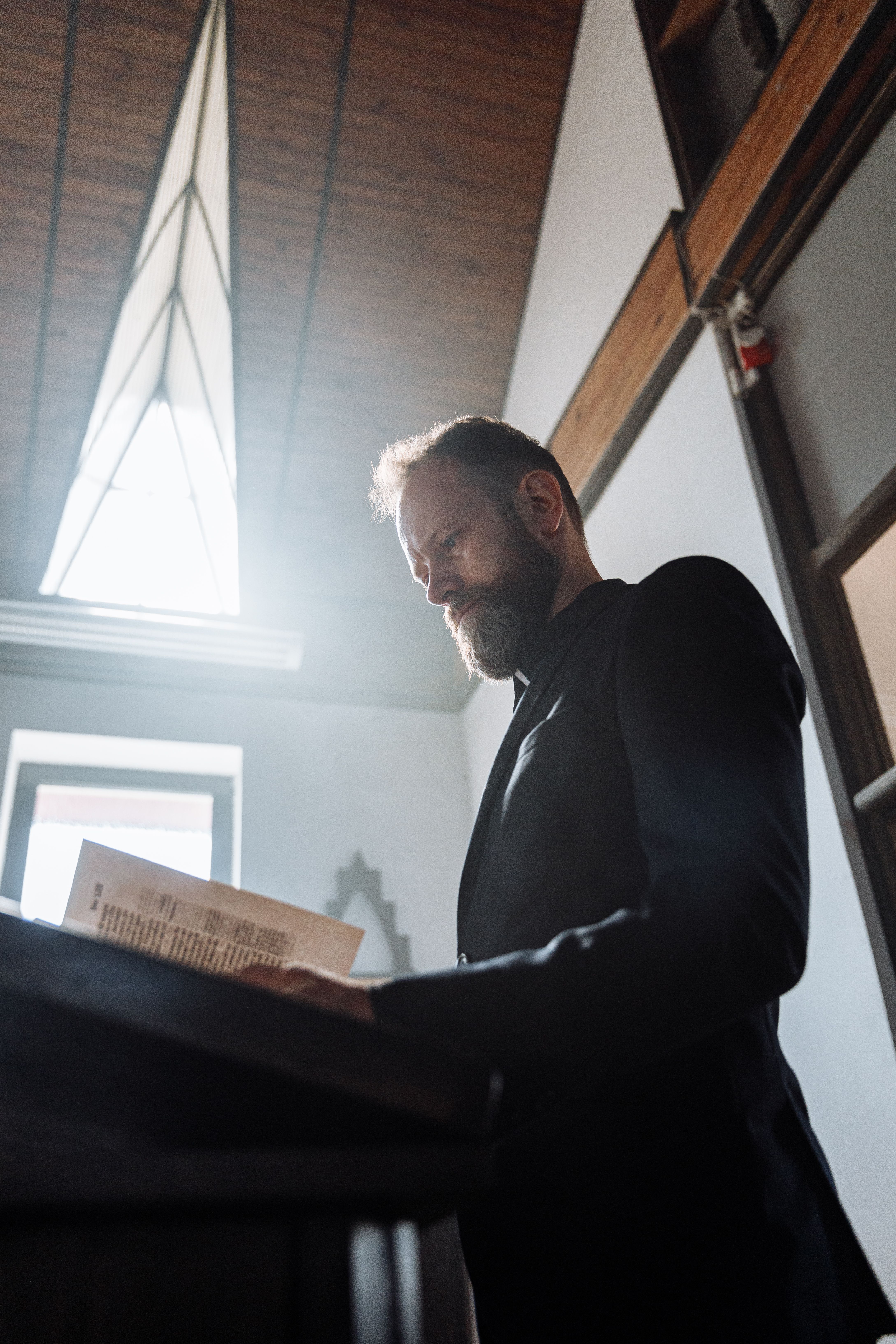 A bearded priest reads the Bible in a sunlit church interior.