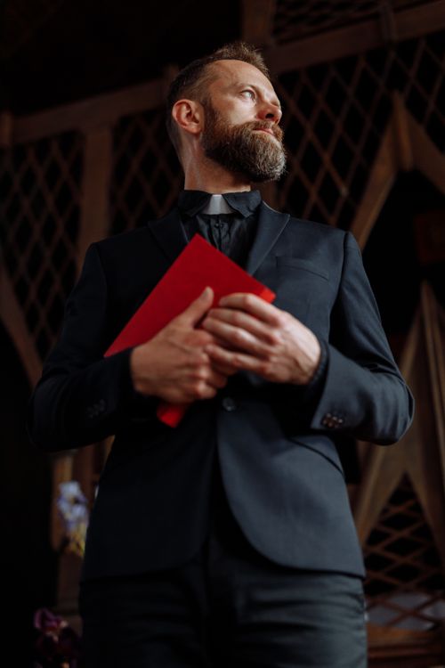 Free Clergyman in traditional attire holding a red book inside a church, conveying a sense of faith and spirituality. Stock Photo