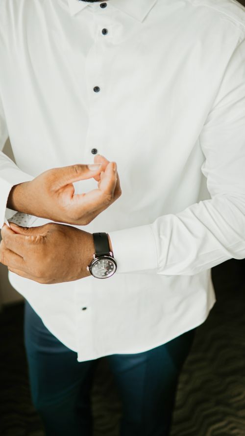 Free Crop anonymous groom with wristwatch buttoning up cuff of classic white shirt while preparing for wedding celebration in light room Stock Photo