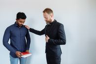 Man with Closed Eyes Holding a Red Book While Man in Black Suit Holding Hand on His Shoulder