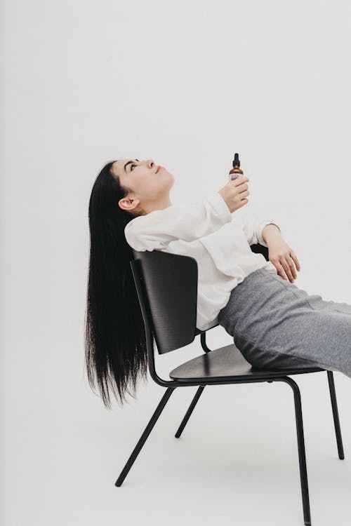Free A Woman in White Long Sleeves Sitting on the Chair while Holding a Glass Bottle Stock Photo