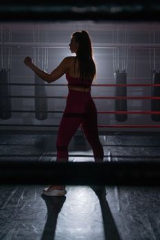 Powerful silhouette of a woman practicing boxing in a dimly lit ring.