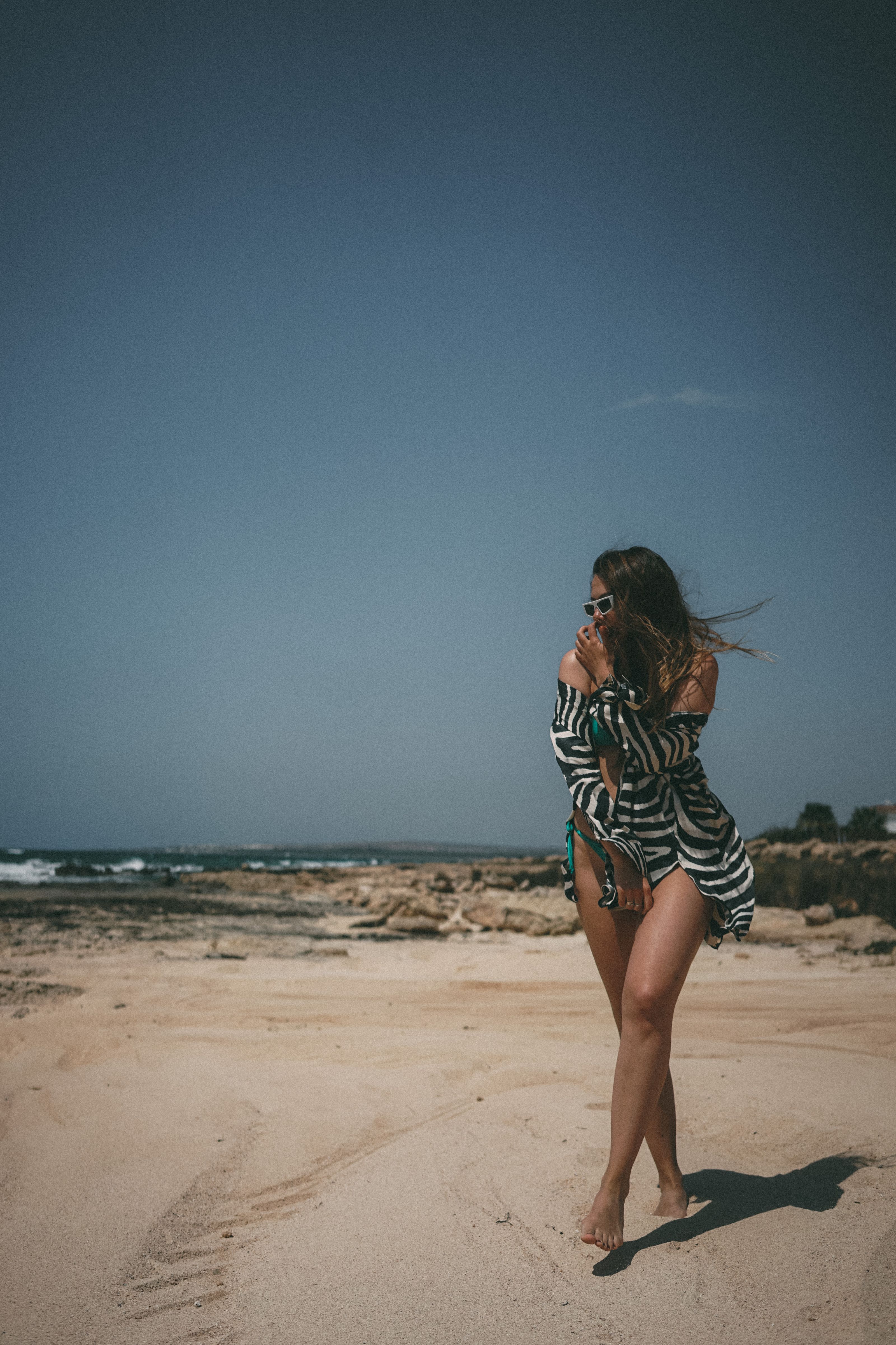 Uma mulher em um vestido listrado e óculos de sol aproveitando um dia de verão ventoso na praia.