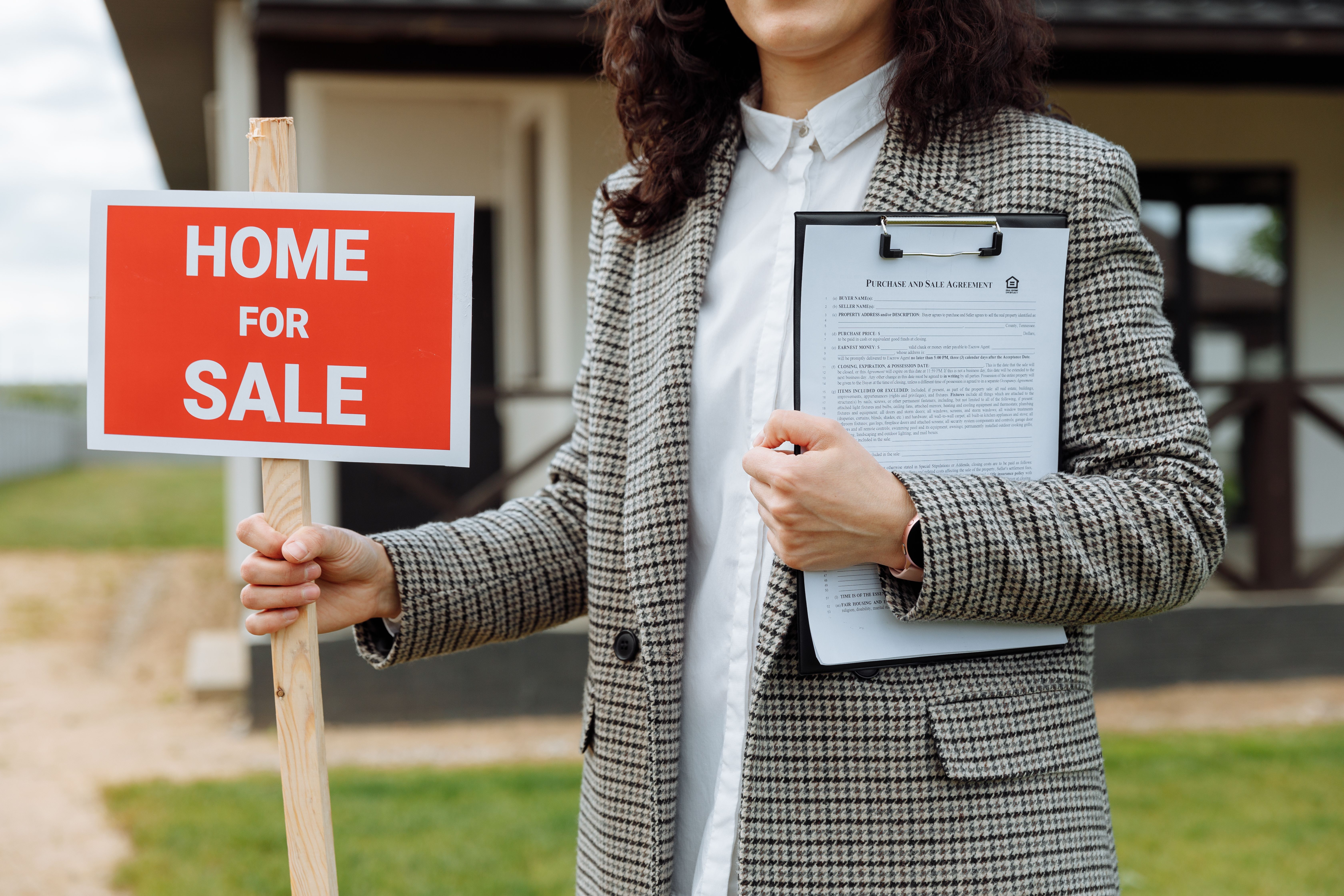 Alt Text: A real estate agent holding a white and red "Home for Sale" sign and a clipboard, standing outside a brick residential property.