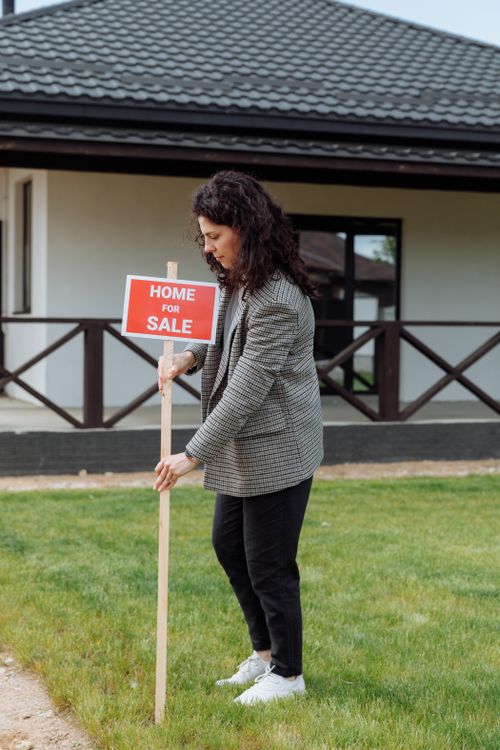 Free Woman placing a home for sale sign in front of a house, showcasing real estate career. Stock Photo
