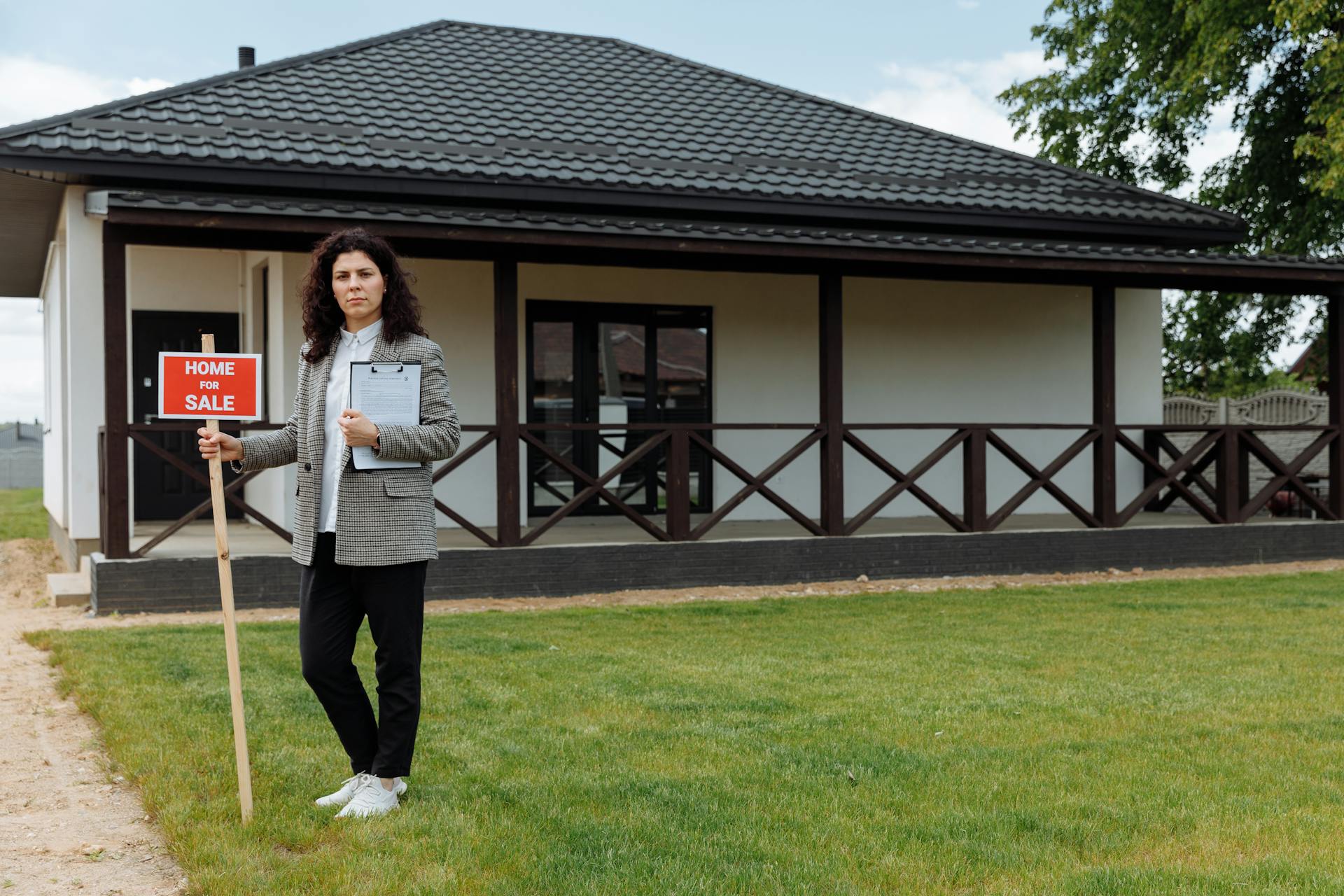 Real estate agent standing outside a house with a 'Home for Sale' sign on a grassy lawn.