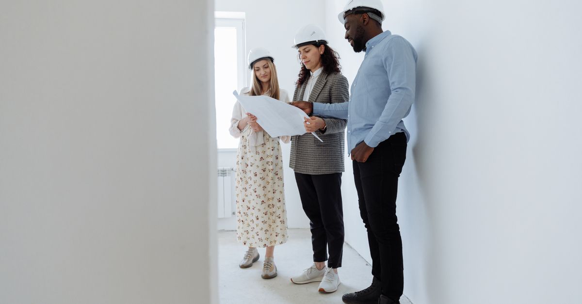 Three architects in hard hats review blueprints in a bright, empty white room.