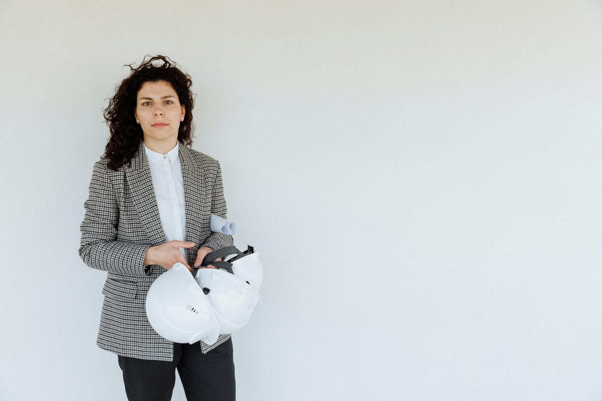 Confident female architect holding a hard hat and architectural plans against a white wall.