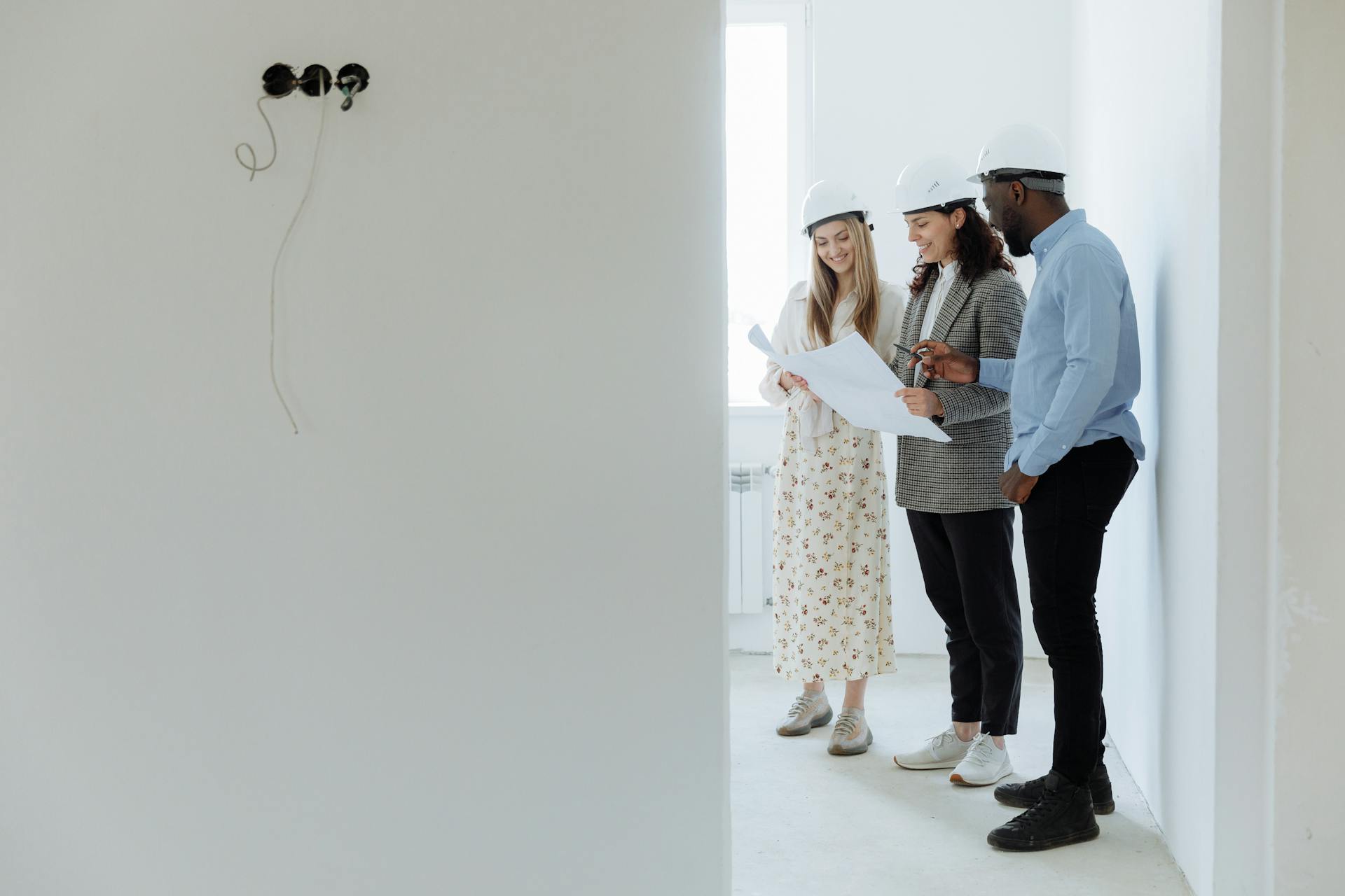 Three architects with hard hats reviewing building plans in an unfinished room.