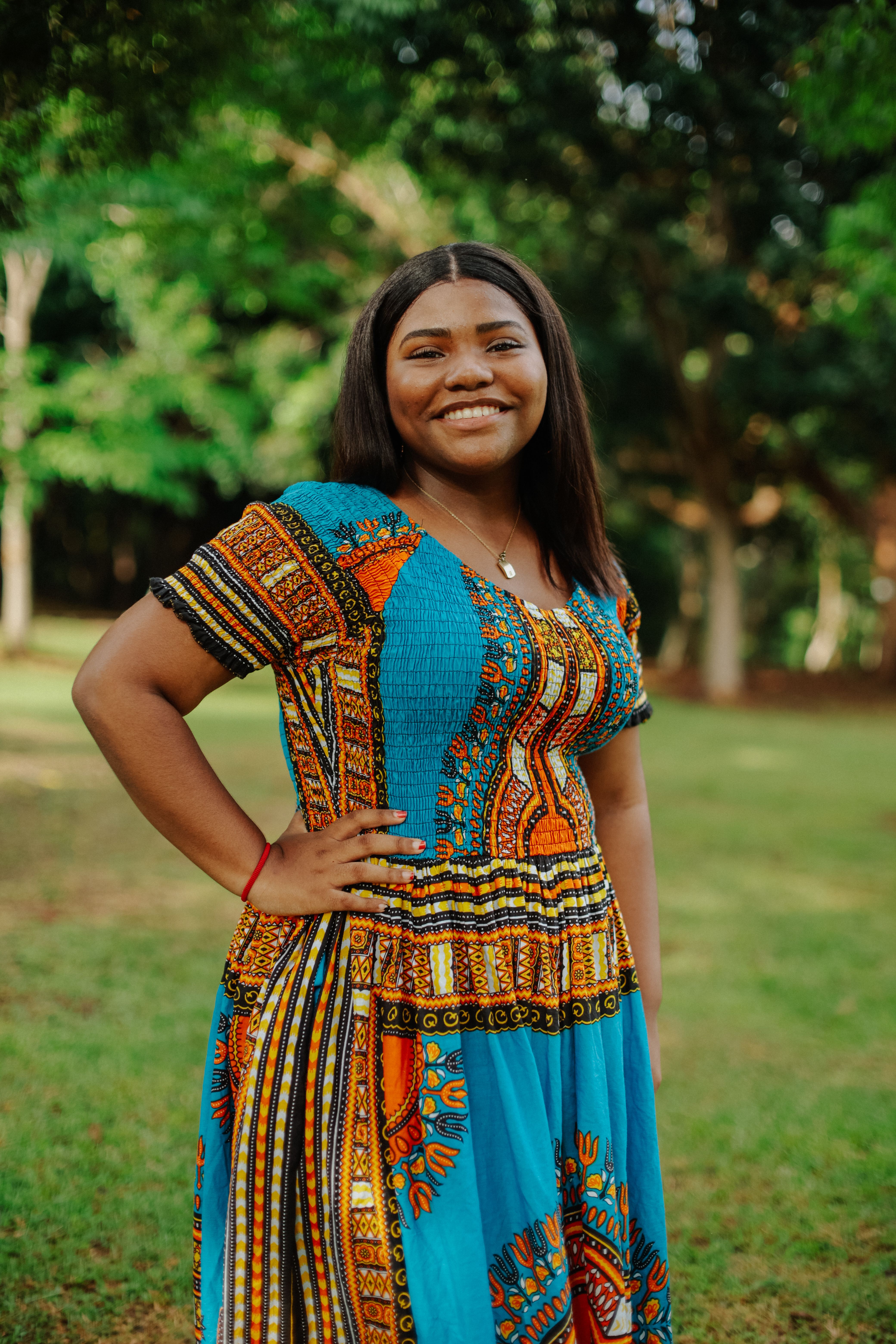 Happy African woman smiling with hand on hip in a vibrant park setting, wearing a colorful dress.