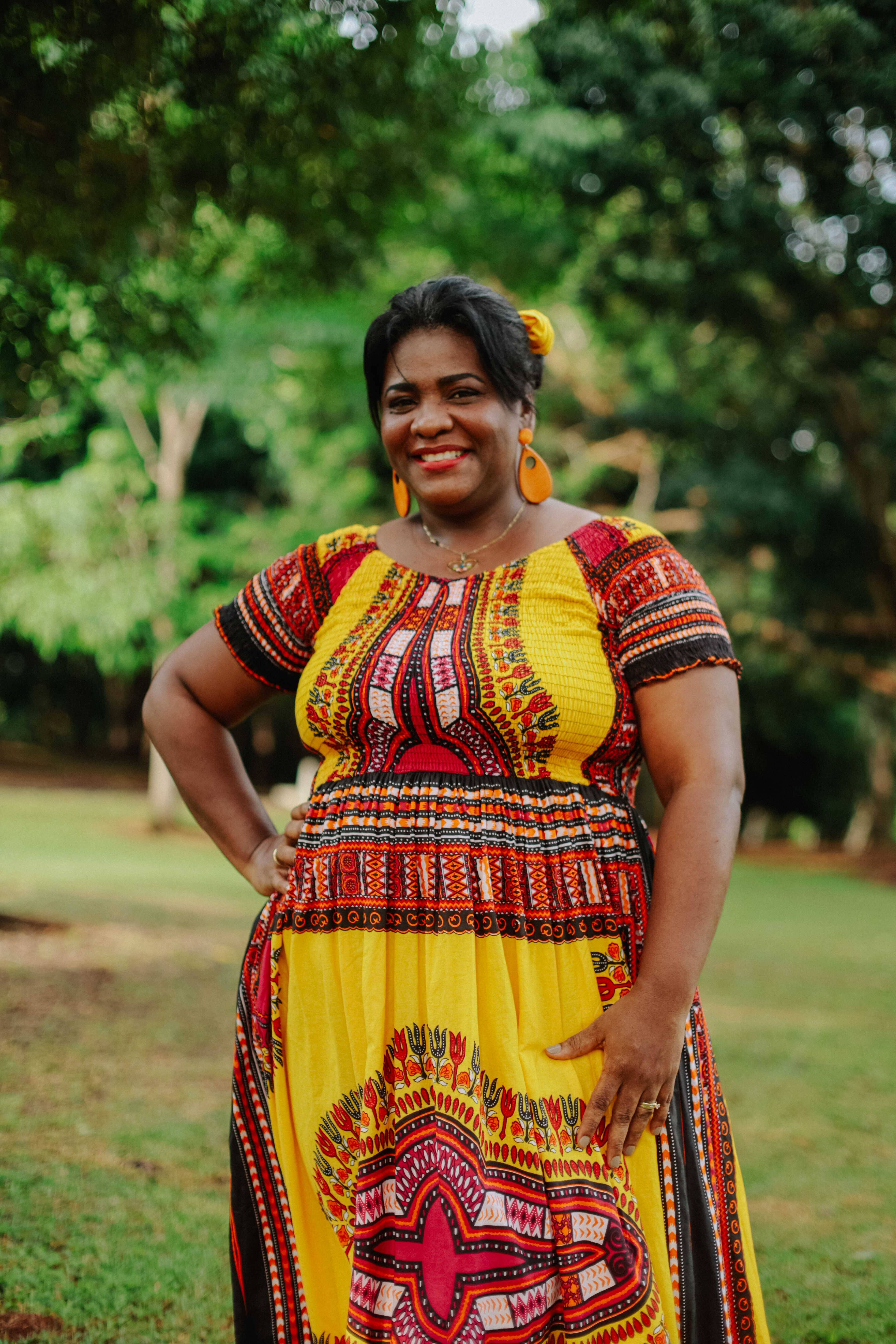 A smiling woman in a colorful dress posing outdoors surrounded by greenery.