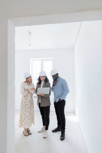 Three architects in a minimalist room discussing plans with a laptop wearing helmets.