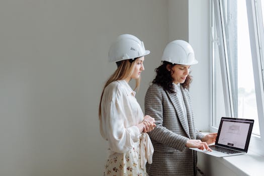 Two female professionals in hard hats review construction plans on a laptop by a window.