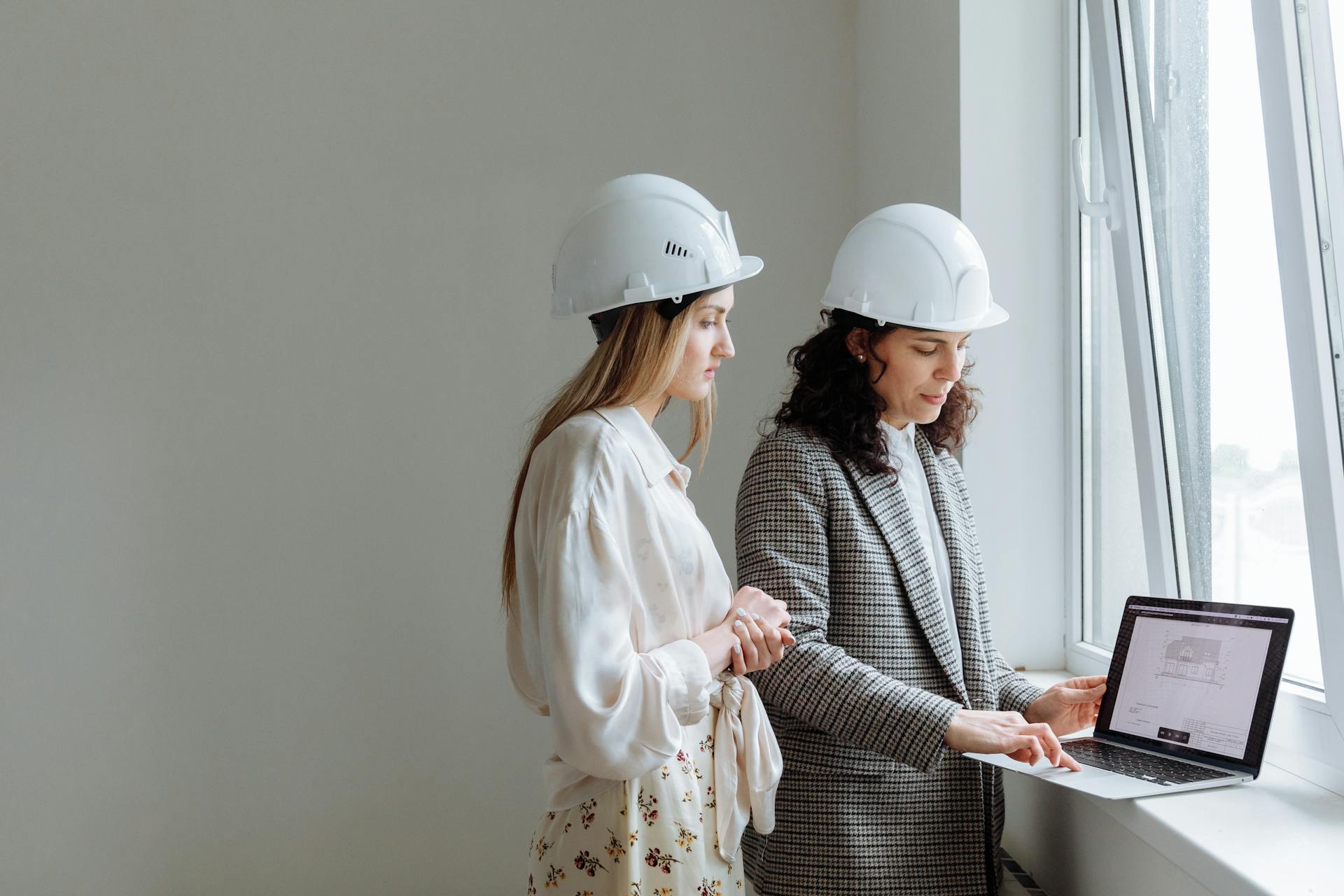 Two female professionals in hard hats review construction plans on a laptop by a window.