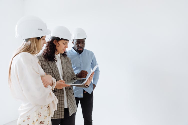 People Wearing Hard Hats Standing Near White Wall While Looking At The Screen Of Laptop