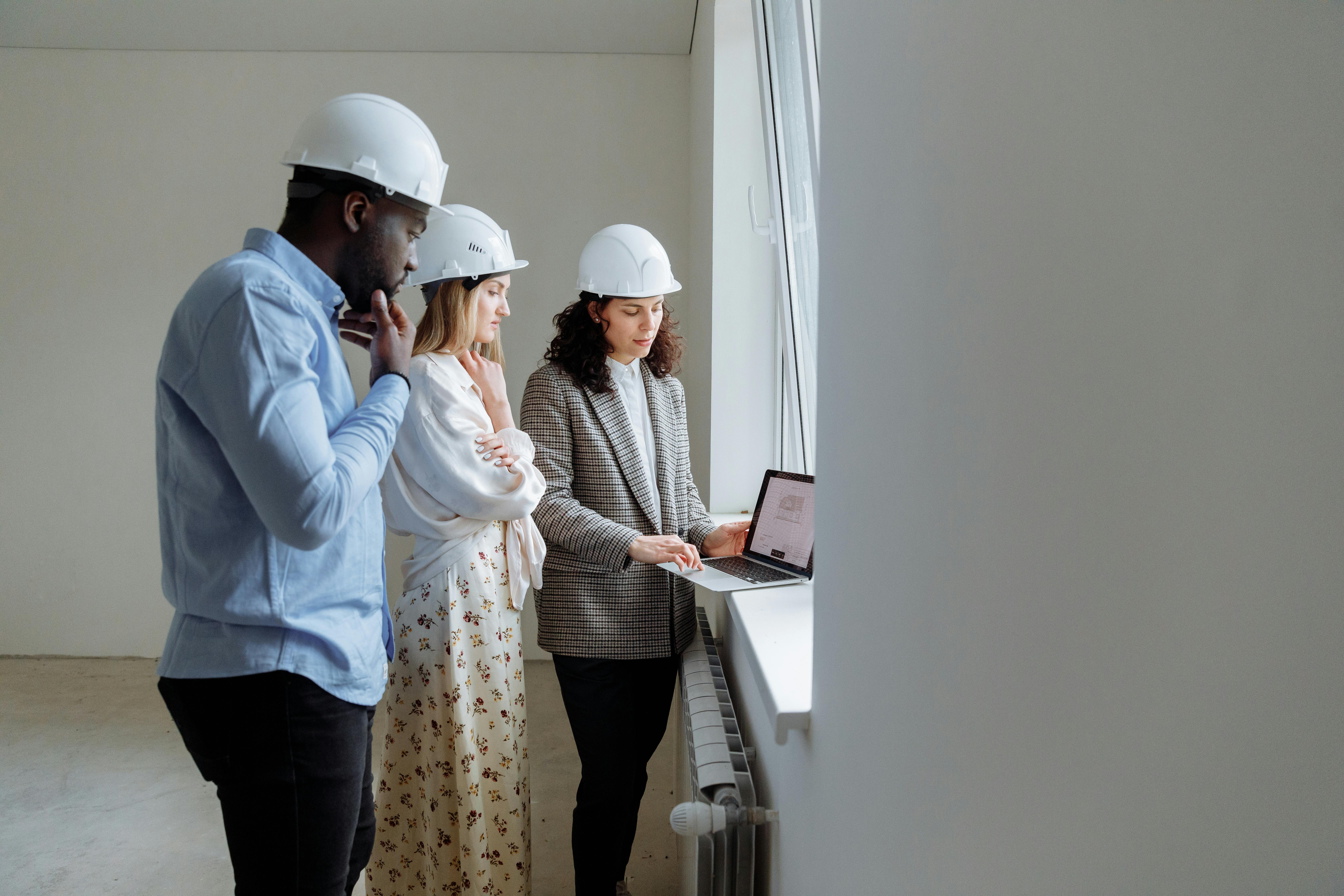 Three engineers wearing helmets review building plans on a laptop in a bright indoor setting.