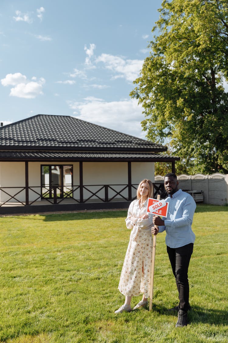 Man And Woman Standing On The Front Yard Holding A Plate Saying That The House Is Sold 