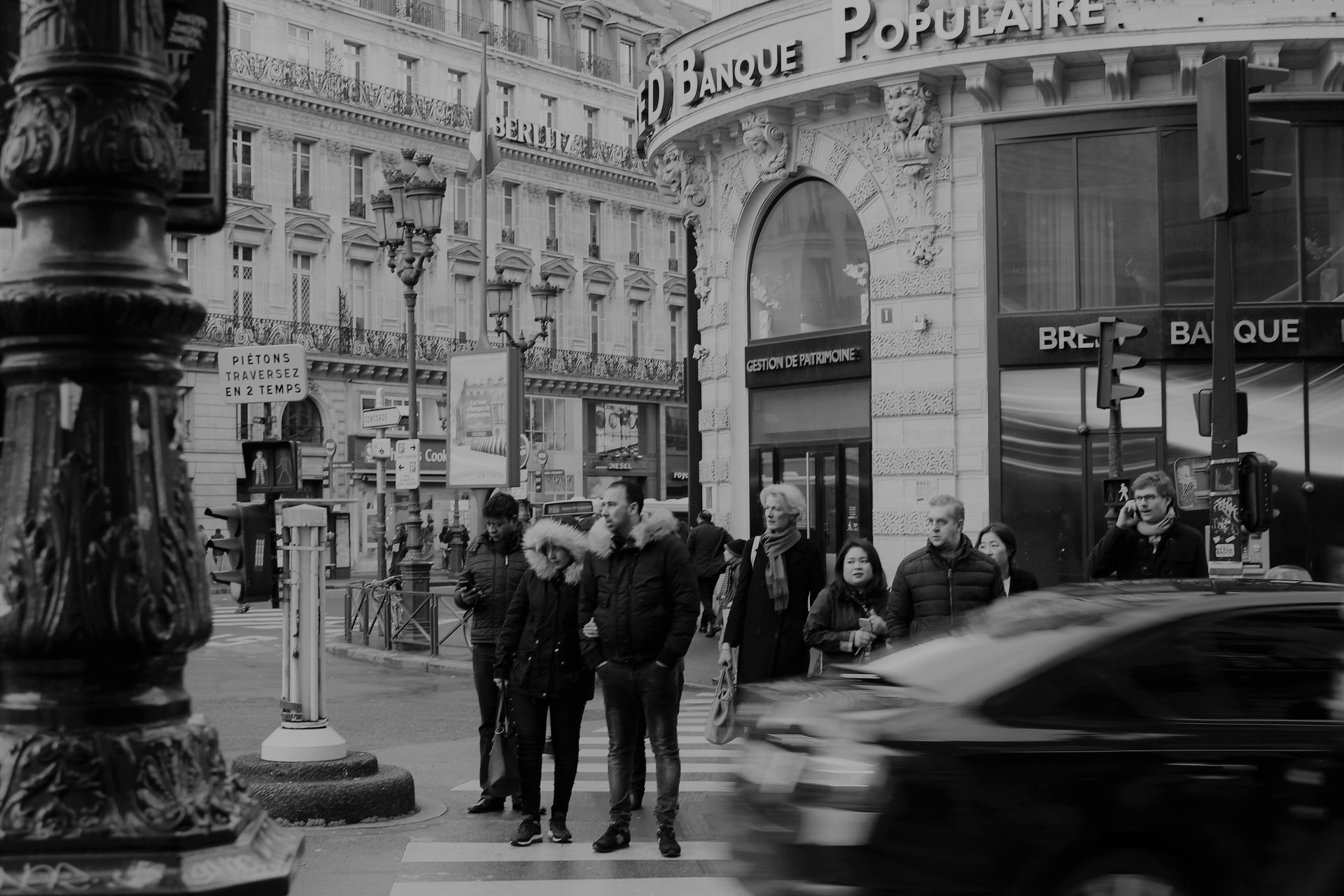 Monochrome view of people crossing a street in front of a classic Parisian building.