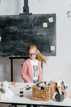 A young girl wearing glasses and a cardigan stands in a classroom near a chalkboard, showcasing creative expression.