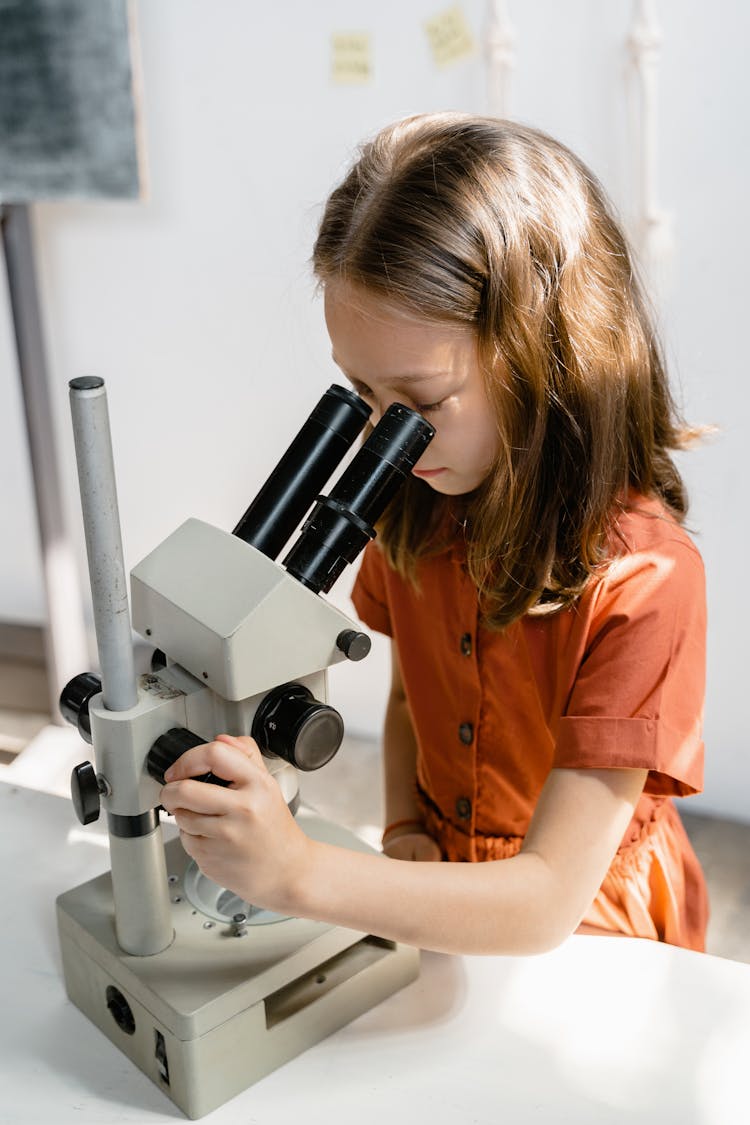 A Girl In Orange Dress Using The Microscope On The Table