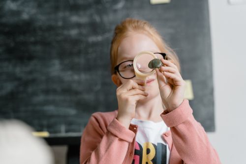 Free Young girl in classroom scrutinizing a rock using a magnifying glass, showcasing curiosity and science. Stock Photo