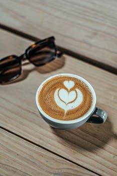 A beautifully crafted latte with latte art on a wooden table beside sunglasses.