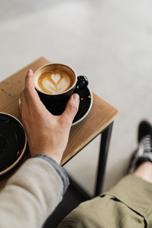 Free Close-up of a hand holding a latte with heart art in a black cup on a saucer. Stock Photo