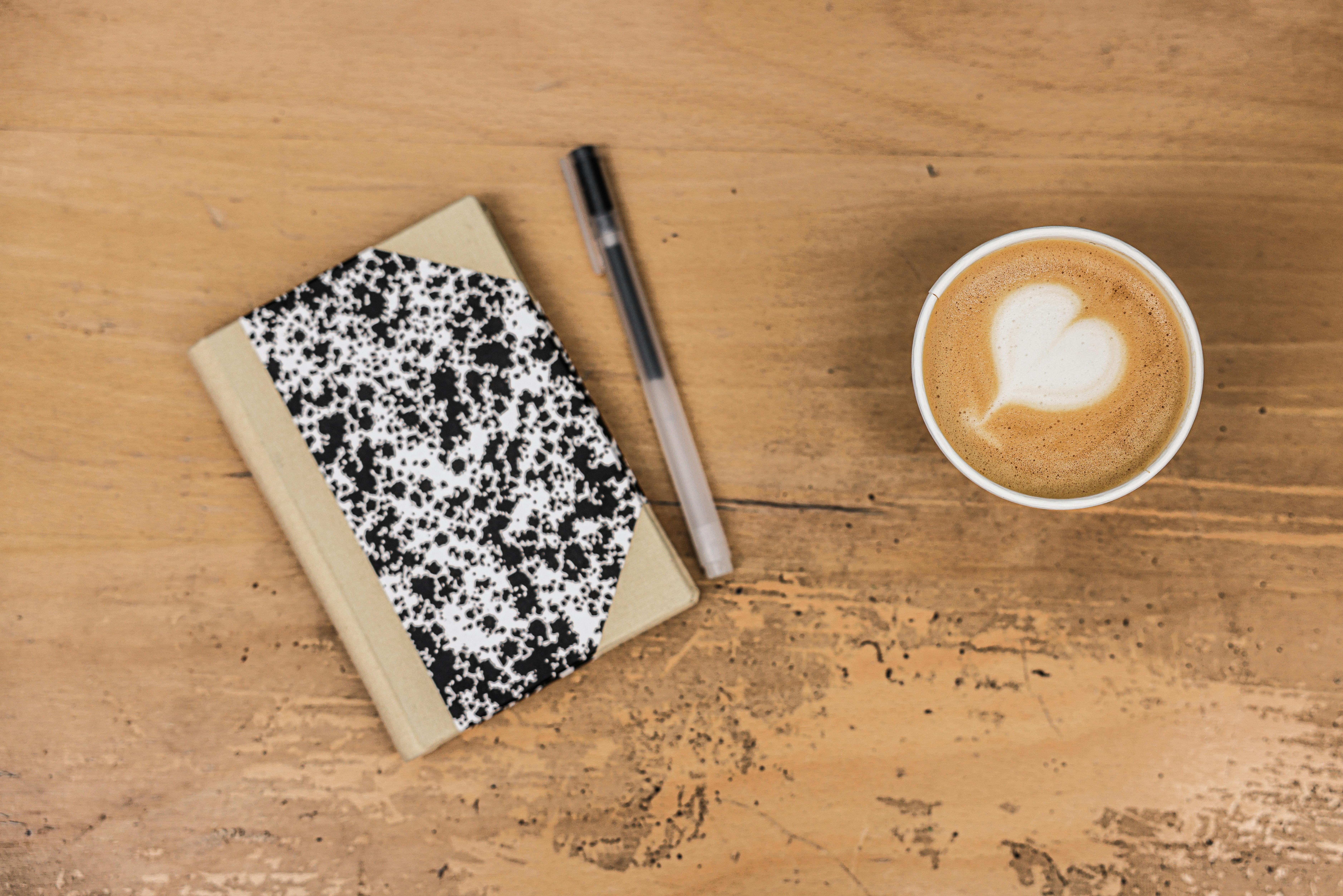 A comforting flatlay featuring a latte, marble notebook, and pen on a wooden table.