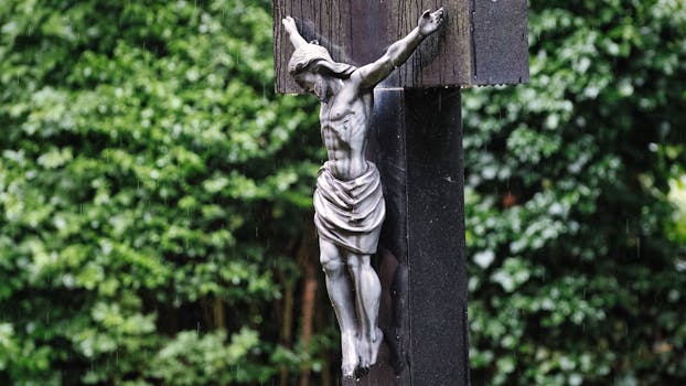 A close-up of a wooden crucifix depicting Jesus, set against a green garden background under rain.