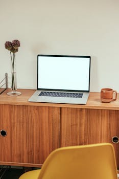 A wooden desk with an open laptop, vase with flowers, and a mug in a minimalist home office.