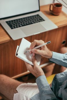 Close-up of hands writing in a notepad at a desk with a laptop, suggesting a work-from-home setup.