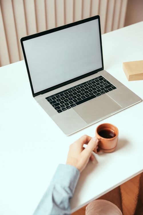 Free A clean workspace featuring a laptop and a hand holding a coffee cup, ideal for remote work settings. Stock Photo