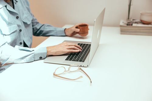 Free Casual workspace featuring laptop, coffee cup, and eyeglasses on a white table. Stock Photo